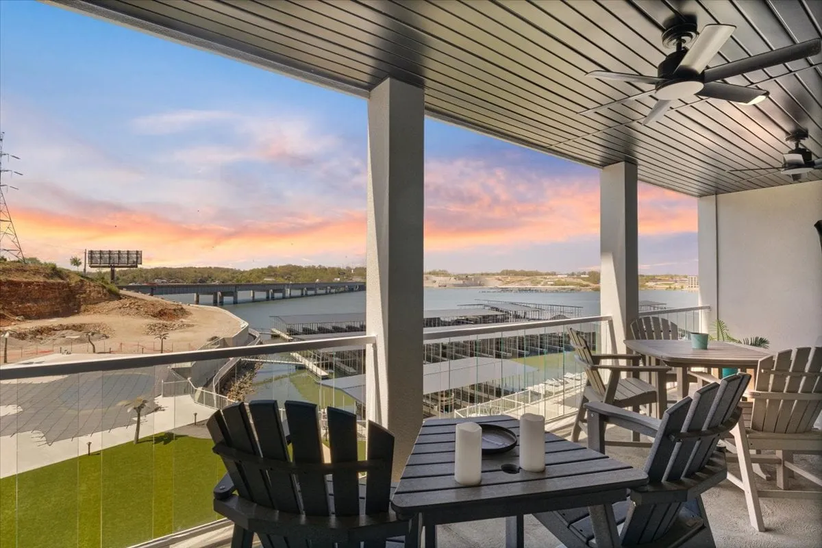 The image shows a serene balcony at Lookout Lodge with comfortable seating overlooking the sparkling waters of Lake of the Ozarks. The scene is peaceful, with the expansive lake stretching out toward a distant tree-lined shore—perfect for sunset watching, gathering with friends, or simply unwinding after a day of lake adventures.
