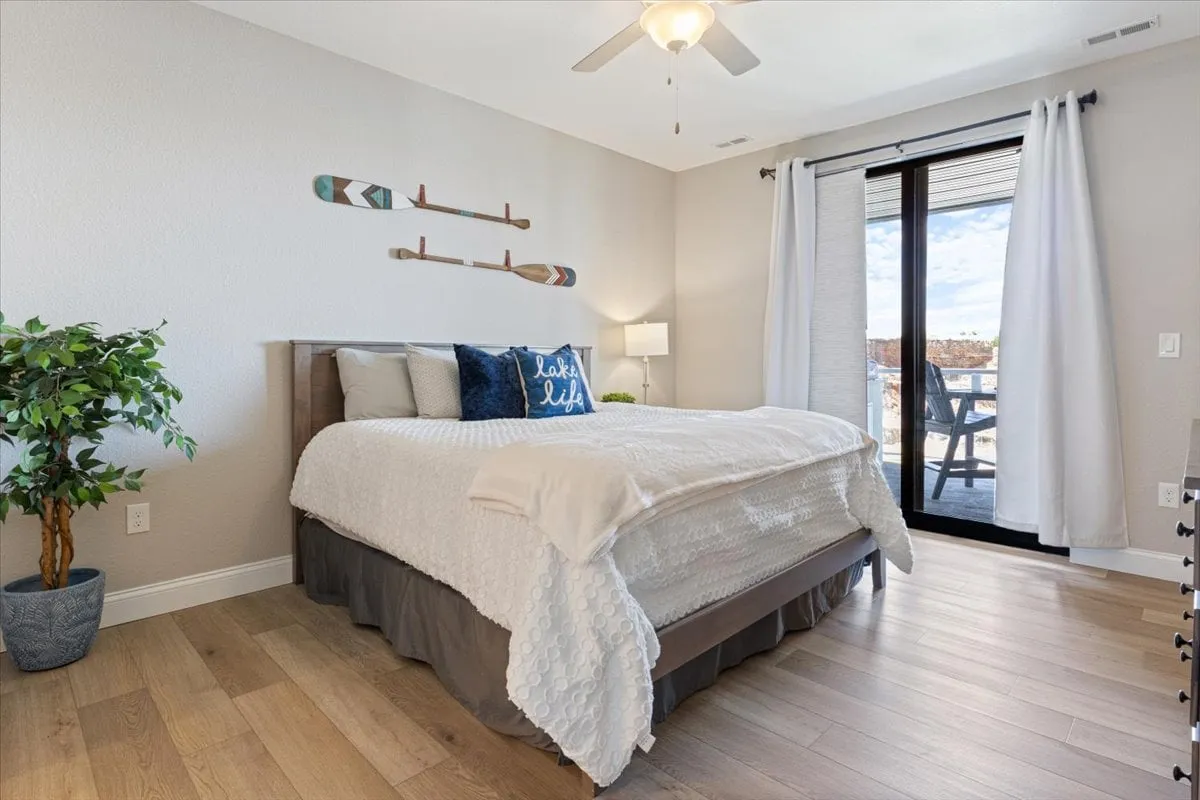 A beautifully appointed bedroom with a plush bed adorned with decorative pillows, a nightstand with a lamp for reading, sliding glass doors leading to a balcony, decorative wall hangings, and an indoor plant. Natural light softly fills the room, suggesting the possibility of sunrise views.
