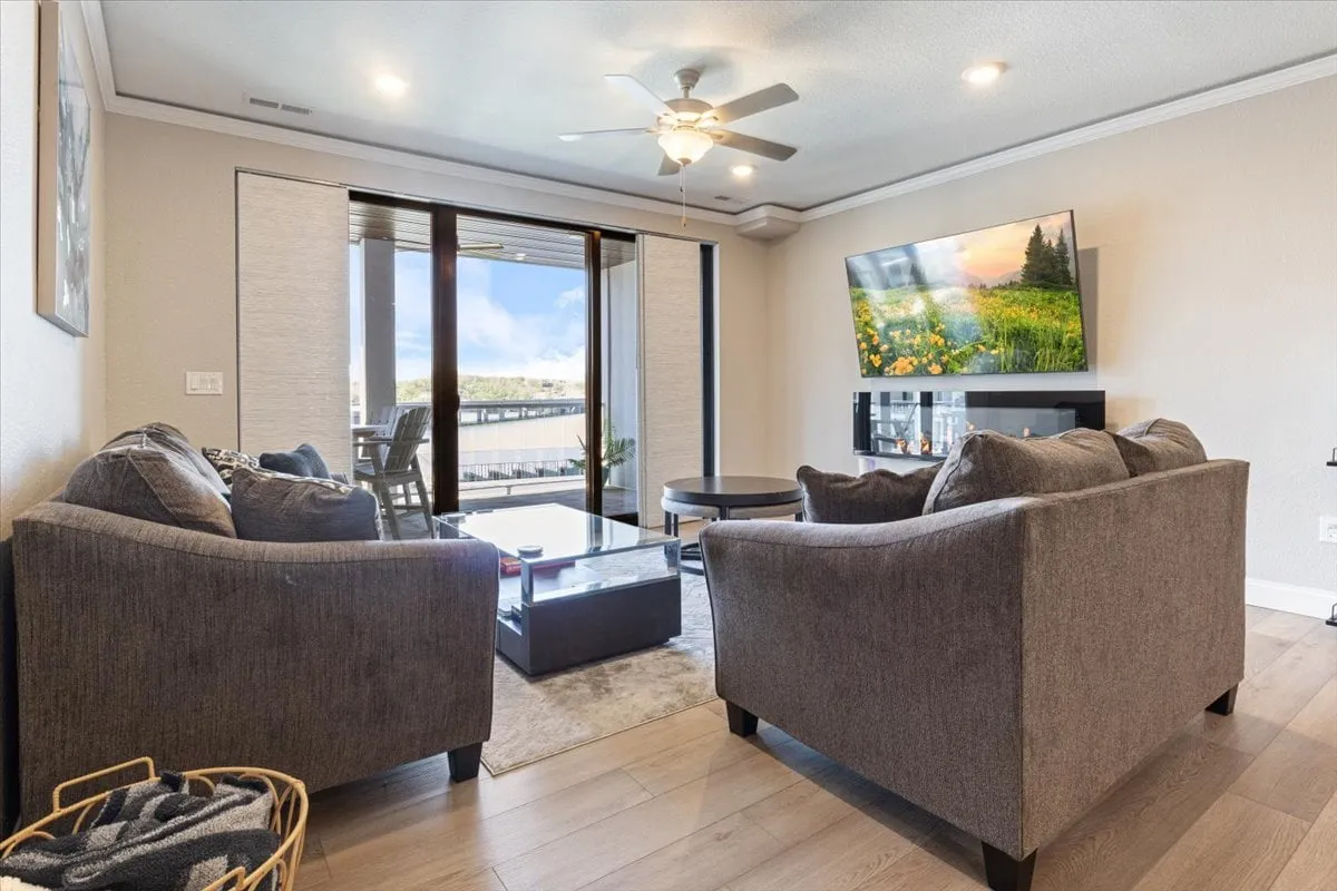 A spacious living room furnished with comfortable seating, a large television, and expansive windows that flood the space with natural light and frame breathtaking main-channel lake views. The setting is ideal for relaxing, entertaining guests, or enjoying a movie night, with the lake's sunset visible through the windows.