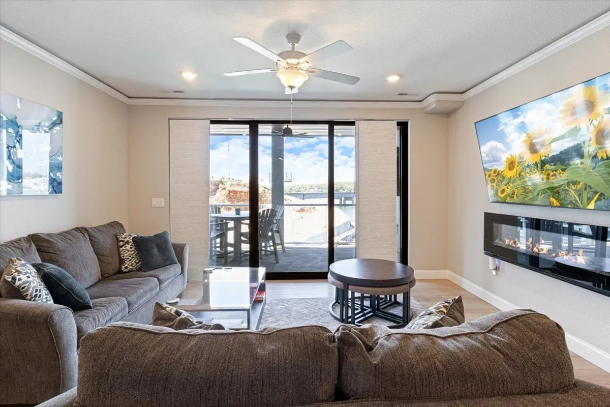 A cozy living area with a comfortable gray sofa and modern coffee table, positioned in front of large windows that offer a sweeping view outside. A vibrant sunflower artwork hangs on the wall, and a fireplace promises warmth and relaxation. The space feels inviting for morning coffee or evening unwinding while enjoying the scenery.