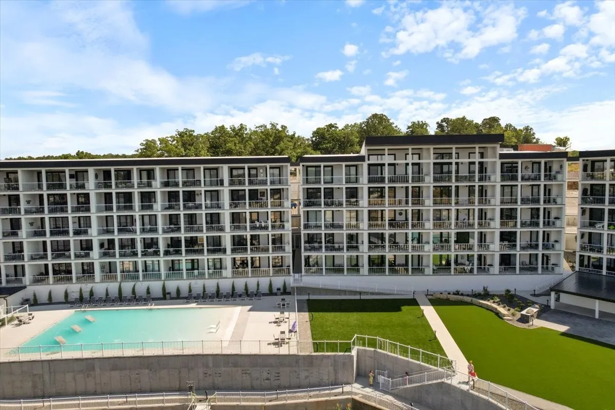 A vibrant exterior setting with a swimming pool, green lawn, balconies, and surrounding trees. The image evokes leisure and sociability with people swimming, sunbathing, and relaxing outdoors.