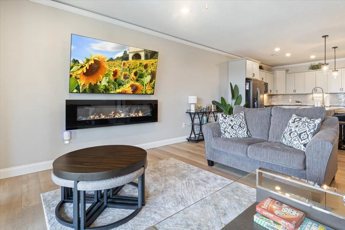 A spacious living area featuring a large TV displaying a sunflower field, a modern electric fireplace, a comfortable grey sofa with decorative pillows, and a stylish coffee table. The room exudes warmth and is perfect for movie nights, reading, or gathering with friends and family during any season.