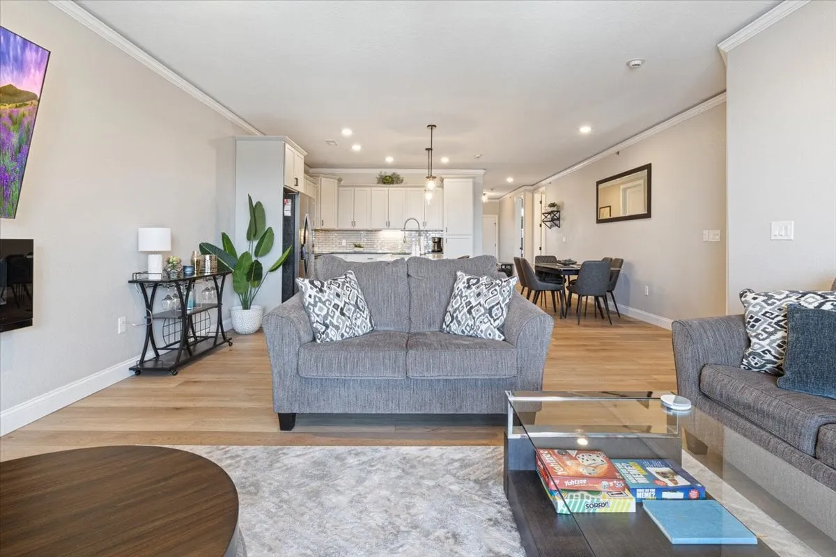 A comfortable living space with a plush sofa, decorative pillows, a coffee table set with board games, and the kitchen visible in the background. Wall art adds a welcoming touch, and the setup invites family game nights and casual gatherings.