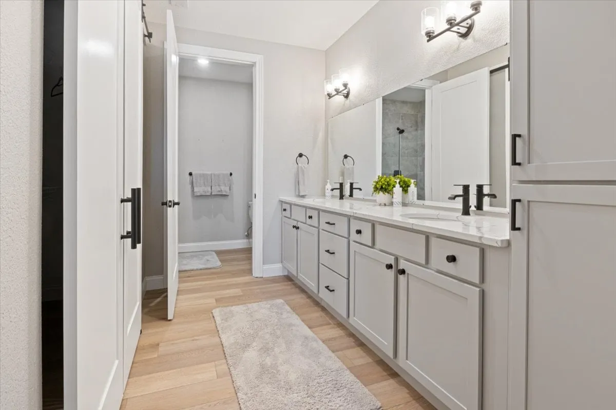 A modern bathroom with a spacious double vanity, a large mirror, elegant light fixtures, and wooden flooring. The space feels calm, organized, and bright—ideal for morning routines and self-care.